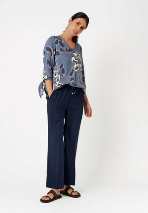 Frau steht mit den Händen in den Taschen, trägt eine blaue geblümte Bluse, marineblaue weite Hosen und braune Sandalen vor einem einfarbigen weißen Hintergrund.