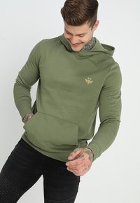 Hoodie vert olive avec un emblème doré, manches longues et poche kangourou. Fabriqué en matériau doux avec une coupe décontractée, comprenant des poignets en côtes.