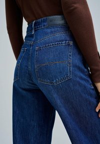Jeans de mezclilla azul de tiro alto con un lavado sutil, costuras visibles y dos bolsillos traseros. Etiqueta de marca "SALSA JEANS" en la cintura.