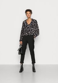 Blusa preta com padrão floral, decote em V profundo e mangas bufantes, combinada com calças pretas ajustadas e botins pretos. Segurando uma pequena bolsa preta.