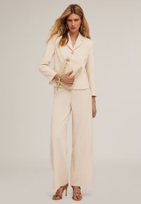 Blazer moulant crème avec un bouton unique, pantalon large crème et talons. Tiens une pochette texturée crème avec un accent en chaîne.