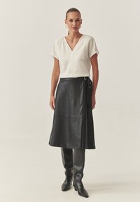 Schwarzer Lederwickelrock mit seitlichen Knopfakzenten, kombiniert mit einer weißen V-Ausschnitt-Bluse. Das Outfit wird von eleganten, figurbetonten schwarzen Stiefeln bis zu den Knien ergänzt.