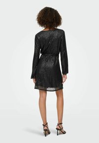 Crna haljina s pailettes dugih rukava, remenom u struku i dužinom do koljena. Ima glatku teksturu i sjajni uzorak. Uparena s crnim sandalama na visoku petu.