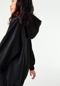 Černá oversized mikina s kapucí na stažení, žebrovanými manžetami a měkkou, strukturovanou tkaninou, zobrazená z boku na světlém pozadí.