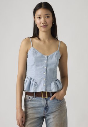 Mladá žena s dlhými tmavými vlasmi nosí svetlomodrý bezrukávový top na gombíky a svetlomodré džínsy s hnedým opaskom, stojí s rukou v vrecku.