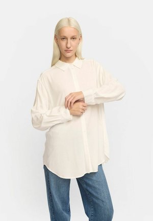 Witte langslurf shirt met een kraag, gladde textuur, knopen aan de voorkant en een afgeronde zoom. Gecombineerd met blauwe denim jeans.