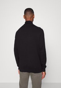 Maglione nero a maglia con colletto alto, polsini e orlo a coste. Presenta una texture liscia con dettagli minimi. Indossato con pantaloni grigio chiaro.