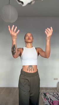 Sieviete ar tetovējumiem uz rokas un vēdera, tērpusies baltā īsajā topā un zaļos biksēs, met baltu priekšmetu telpās pret vienkāršu sienu.