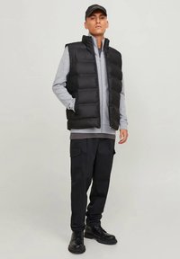 Melna pūkaina veste uz pelēkas rāvējslēdzēja sporta krekla, kopā ar melnām kravas biksēm un melniem zābakiem. Izceļas augsts apkaklis un pūkains dizains.