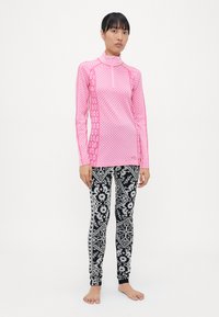 Langermet rosa topp med halv glidelås, med et teksturert mønster. Svarte tights med hvite geometriske og blomstrete design. Barføtt stående.