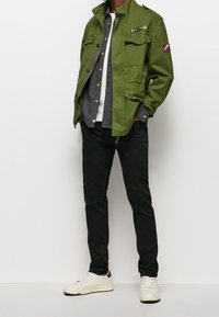 Chaqueta de lona verde con múltiples bolsillos y un logo, superpuesta sobre una camisa gris y una camiseta blanca, acompañada de pantalones negros y zapatillas blancas.