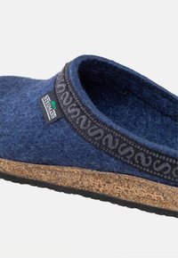 Primo piano di una scarpa blu in feltro senza lacci con bordi neri a motivi, suola in sughero e etichetta del marchio Stichan sul lato.