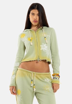 Femme portant un sweat à capuche court vert et un pantalon de survêtement assorti avec des imprimés de soleil jaune et de fleurs blanches, bracelets colorés au poignet droit.