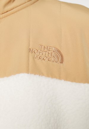 Beżowo-biała kurtka polarowa z wyhaftowanym logo "The North Face" na górnej części klatki piersiowej.