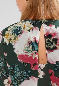Blusa floral de tejido verde con grandes flores rosas y blancas, escote redondo y un pequeño corte en forma de llave en la parte posterior.