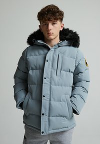 Veste matelassée bleu clair avec capuche bordée de fourrure, boutons noirs et poches latérales ; le motif matelassé crée un effet texturé.