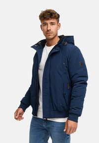 Joven vestido con una chaqueta acolchada azul marino sobre una camisa blanca y jeans azules, de pie contra un fondo claro y liso.