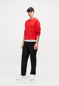 Κόκκινη κουκουλίτσα crewneck με μικρό λογότυπο, φορεμένη πάνω από λευκή μπλούζα, συνδυασμένη με μαύρο τζιν παντελόνι και λευκά αθλητικά παπούτσια. Απλός σχεδιασμός.