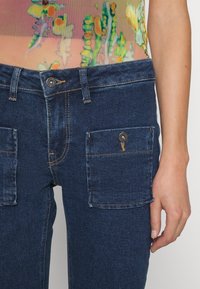 Mörkblå denimjeans med framfickor med klaff och kontrasterande sömmar, bärs med en färgglad semi-genomskinlig topp som visar del av högra handen.