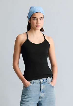 Jeune femme portant un débardeur noir côtelé, un jean bleu clair et un foulard bleu à fleurs sur la tête, debout les mains dans les poches devant un fond uni.