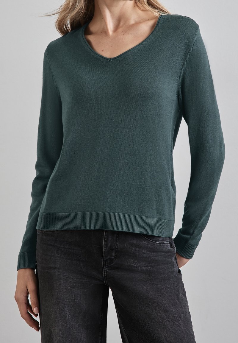 Mörkgrön V-halsad tröja i mjukt stickat material. Har långa ärmar och ribbad fåll, kombinerad med mörka jeans.