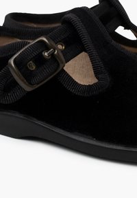 Scarpa in camoscio nero con punta tonda, caratterizzata da una fascia in tessuto texture e un dettaglio in metallo a forma di fibbia. La suola è piatta e realizzata in gomma.