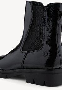 Botte Chelsea noire brillante avec panneau latéral élastique, semelle épaisse et détail de logo rond près de la cheville, photographiée sur fond blanc.
