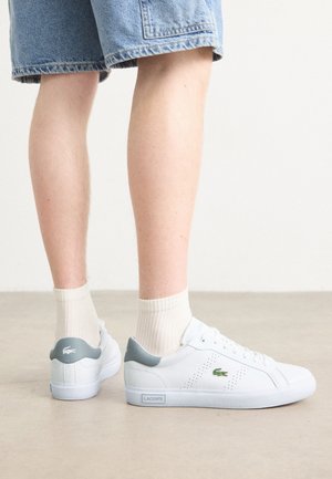 Osoba ubrana w białe niskie trampki Lacoste z zielonym logo, szarą ozdobą na pięcie, białe skarpetki i denimowe szorty stoi na beżowej podłodze.