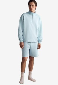 Pulover din fleece de culoare albastru deschis cu guler cu jumătate de fermoar, purtat cu pantaloni scurți asortați. Textură moale, mansete răsucite și croială lejeră. Șosete albe.