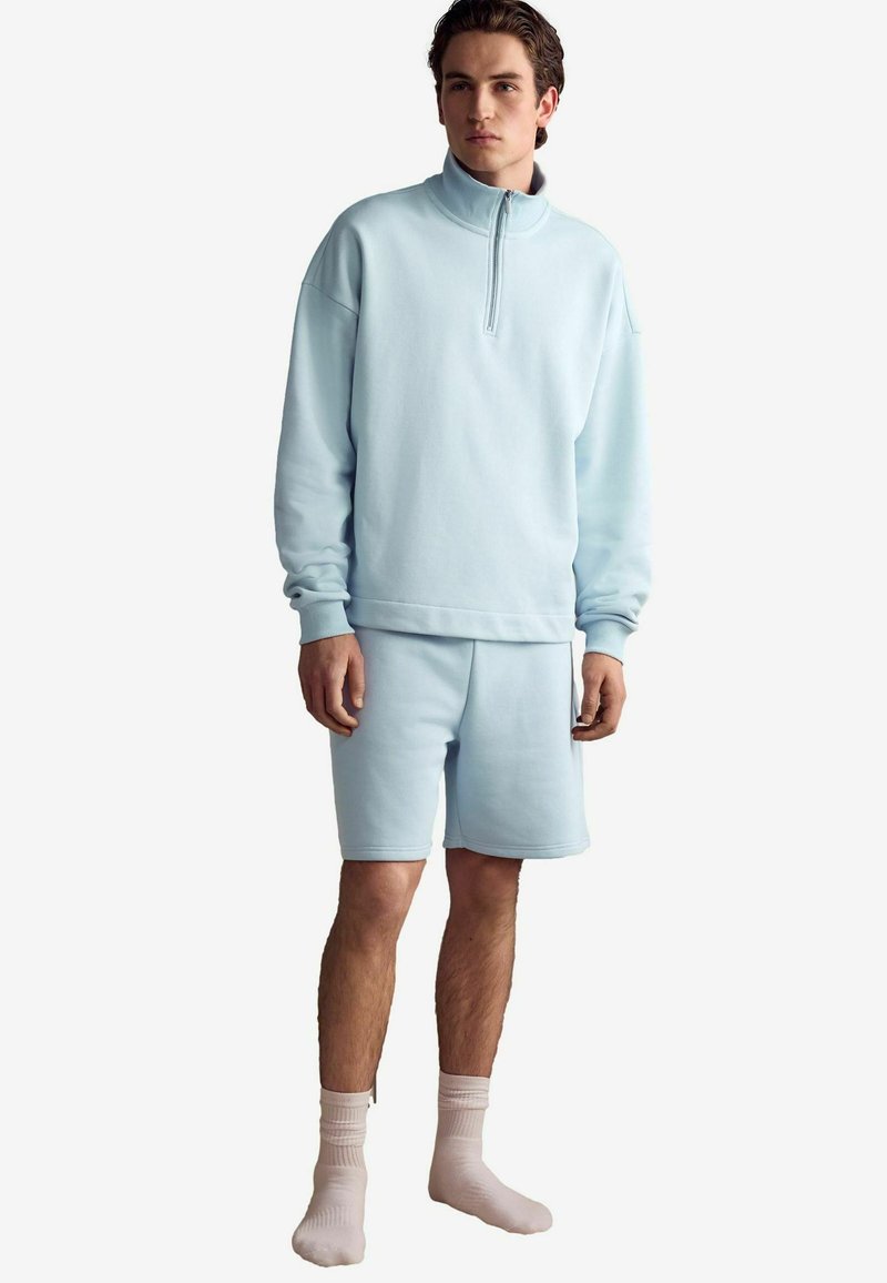 Pulover din fleece de culoare albastru deschis cu guler cu jumătate de fermoar, purtat cu pantaloni scurți asortați. Textură moale, mansete răsucite și croială lejeră. Șosete albe.
