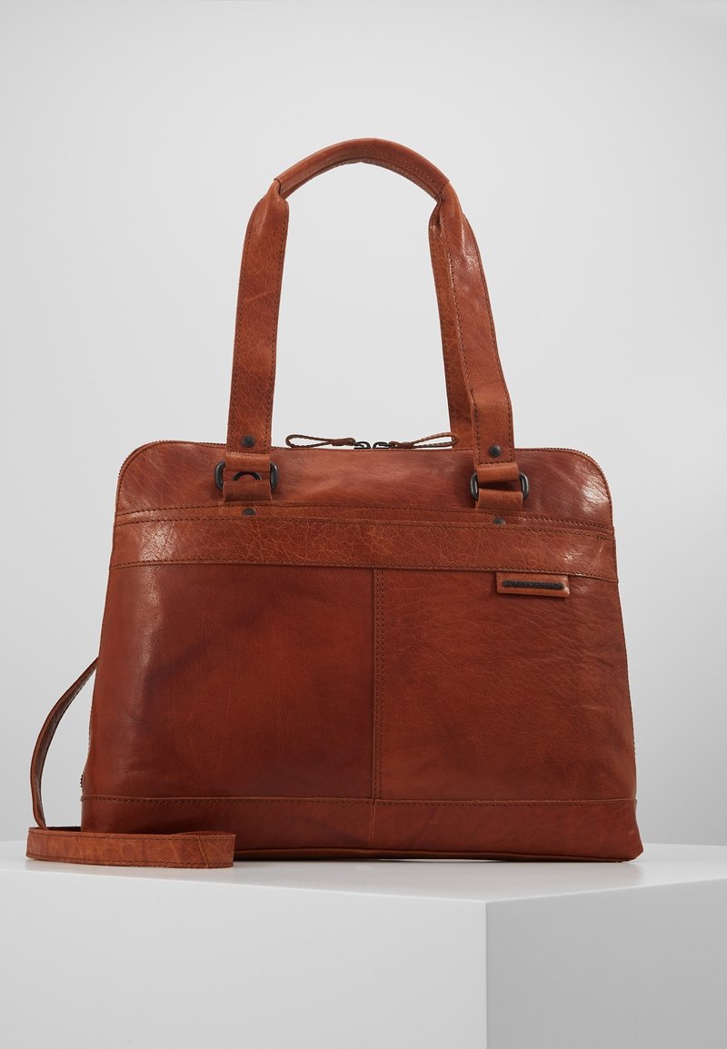 Sac à main en cuir marron avec deux poignées, une bandoulière amovible et une fermeture éclair supérieure, présenté sur un socle blanc devant un fond gris.