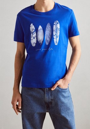 Mężczyzna w niebieskiej koszulce z czterema wzorami desek surfingowych i napisem "wybierz swój styl", w niebieskich dżinsach, z ręką w kieszeni.