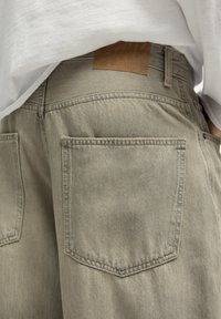 Khaki denim hlače z dvema zadnjima žepoma, teksturirano površino in rjavim usnjenim zaklopom na pasu.