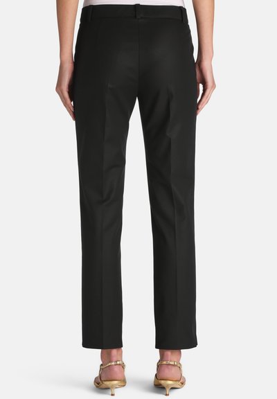 Woman wearing black tailored trousers and gold low-heeled sandals, standing with hands relaxed by sides, shown from the back.