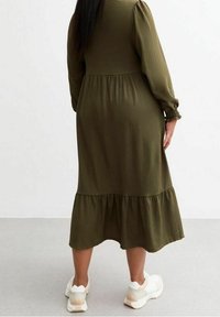 Robe verte olive à manches longues avec un corsage ajusté, des poignets élastiqués et un ourlet à volants en plusieurs niveaux. Portée avec des baskets blanches.