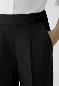 Schwarze, maßgeschneiderte Hose mit strukturierter Oberfläche, breitem elastischem Bund mit "OUi"-Logo und seitlichen Taschen für Funktionalität.