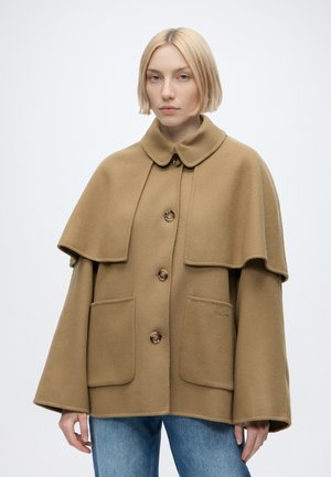 Young woman with short blonde hair wearing a camel-colored buttoned cape coat and blue jeans, standing against a plain white background.