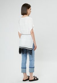 Witte loszittende top met zwarte franjes aan de onderrand en horizontale strepen, gecombineerd met lichtblauwe cropped jeans en zwarte sandalen.