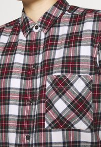 Camisa de cuadros abotonada que presenta cuadros rojos, verdes y negros sobre un fondo blanco. Incluye un bolsillo en el pecho con un patrón de cuadros en diagonal.