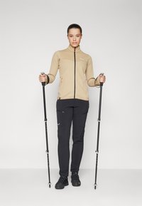 Lys beige, zip-up jakke med teksturert stoff og oransje aksenter, parret med svarte cargo-bukser og turstaver mot en ensfarget bakgrunn.
