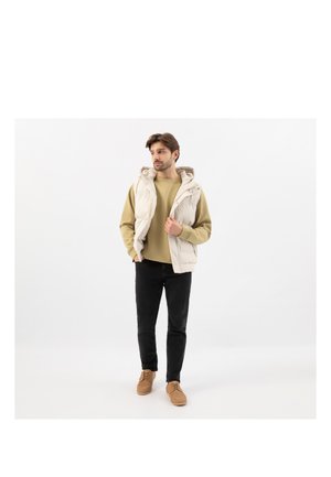 Man die een beige gewatteerd vest met capuchon draagt over een olijfgroene trui, zwarte broek en bruine schoenen, staand tegen een witte achtergrond.