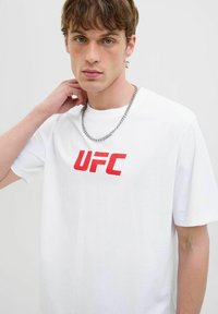 Balta medvilninė marškinėlė, ant kurių priekyje yra ryškiai raudonas "UFC" logotipas, trumpomis rankovėmis, laisvo kirpimo ir apvaliu kaklu. Įtraukta sidabrinė grandinėlė.
