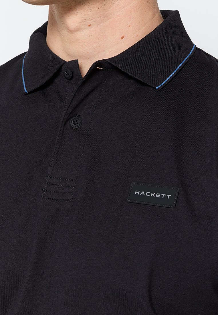 Hombre vistiendo una camiseta polo negra con un cuello de ribete azul y un parche con el logo de Hackett en el pecho.