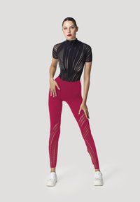 Top in rete nero con maniche corte e colletto alto, abbinato a leggings rosa brillante con ritagli a diamante lungo le gambe. Sneakers bianche.