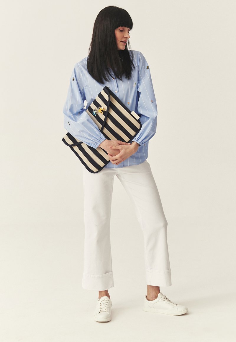 Kobieta w jasnoniebieskiej bluzce i białych spodniach trzymająca dwie czarno-beżowe paskowane kopertówki na jednolitym tle.