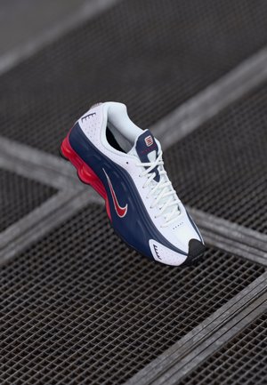 Nike athletic shoe in white, navy, and red. Features a mesh upper, padded collar, and a textured sole. Iconic swoosh logo displayed prominently.