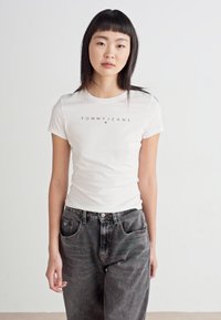 Bílý přiléhavý tričko s logem "TOMMY JEANS" v černé barvě, spárované s šedými džínovými kalhotami s vysokým pasem. Hladký materiál, krátké rukávy, kulatý výstřih.