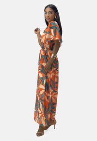 Robe maxi orange avec de grands motifs de feuilles en vert et crème. Manches courtes, taille smockée, tissu léger, associée à des talons beiges.