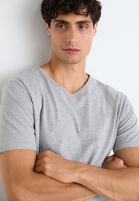 T-shirt gris à col en V avec des manches courtes, présentant une texture douce. Le mannequin se tient les bras croisés devant un fond blanc uni.