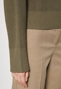 Pull en maille vert olive avec poignets côtelés, associé à un pantalon beige sur mesure. Le pull présente un motif tissé texturé.
